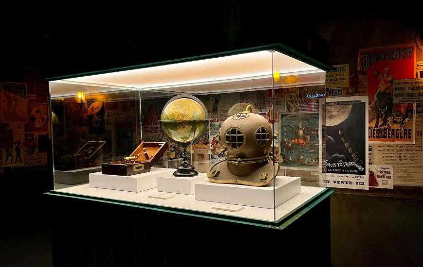 Room 03, The Vernian Work — a display case with an antique diving helmet, a globe and 19th-century instruments, surrounded by vintage posters