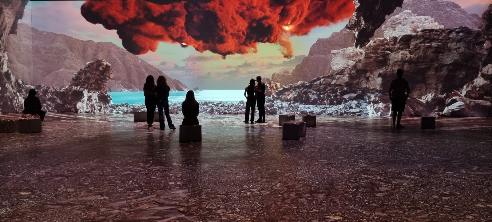Room 04, Le Grand Planisphère Numérique — visitors silhouetted against a floor-to-ceiling projection of a red volcanic cloud over an underground sea